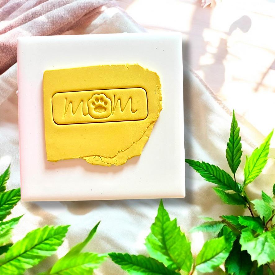 Yellow clay slab with "mom" script and paw print tag impression, set on a white ceramic tile.