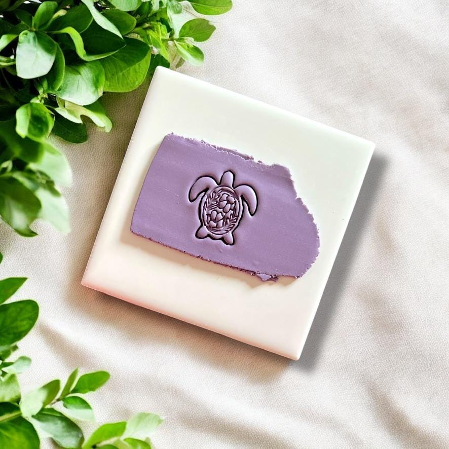 Impression of a folk art turtle design pressed into purple polymer clay with tropical leaves beside