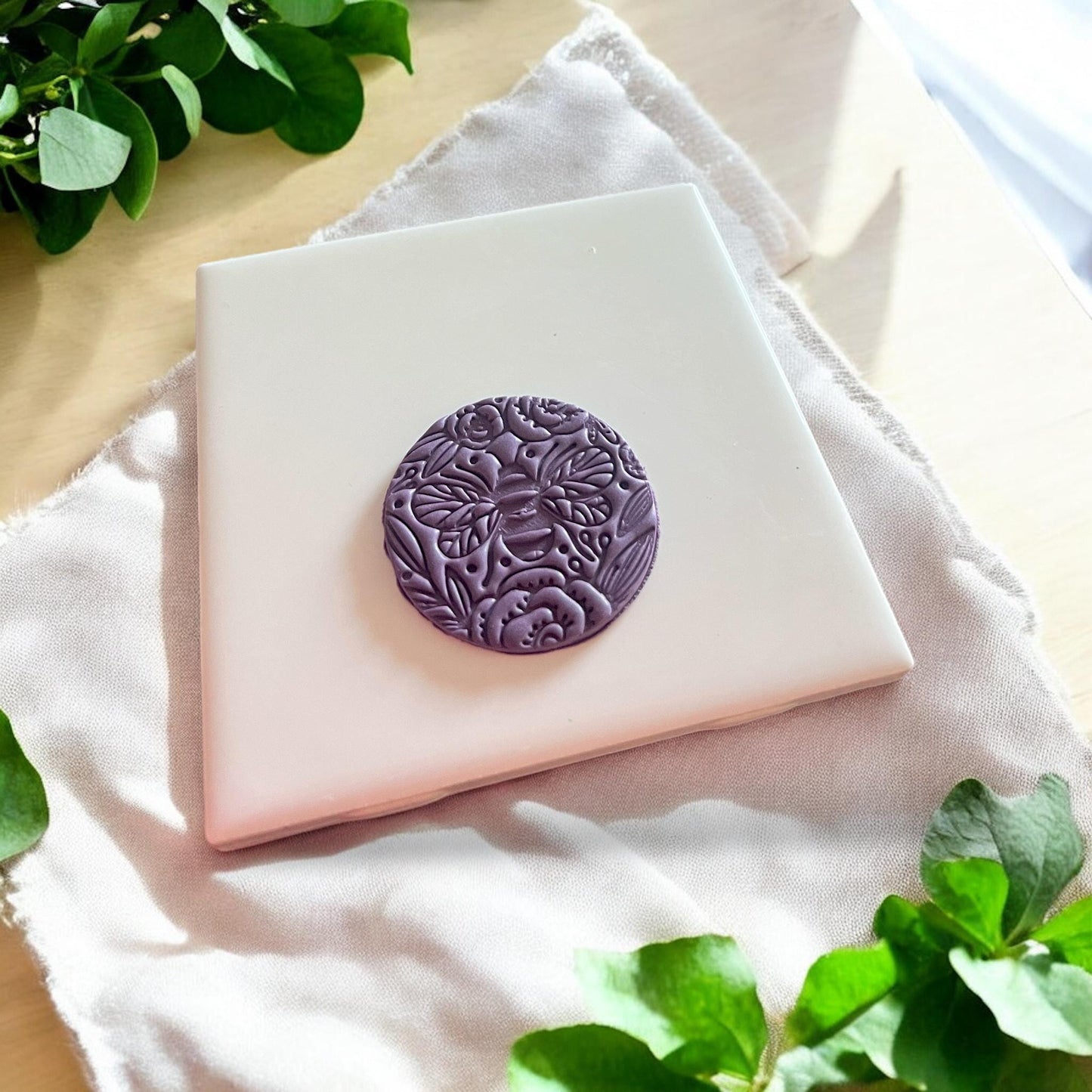 A round purple clay pendant featuring a bee surrounded by floral details, ready for necklace or keychain making