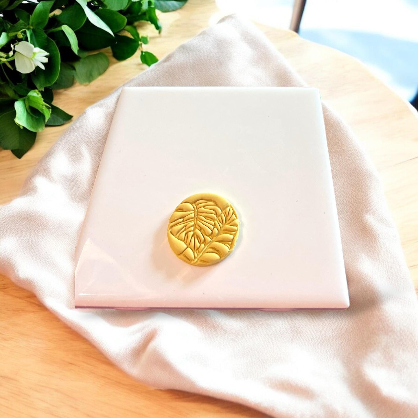 Finished clay piece with tropical leaf design next to the pink circular monstera cutter on a tile.