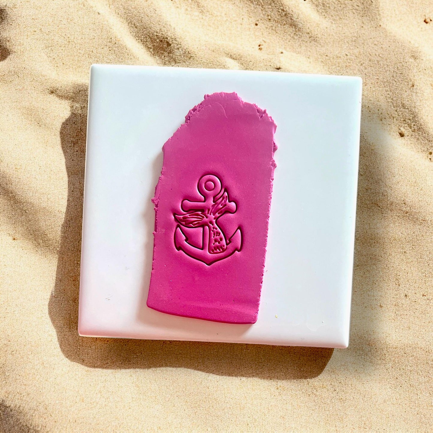 Pink polymer clay impression of mermaid tail anchor on white tile with sand background.