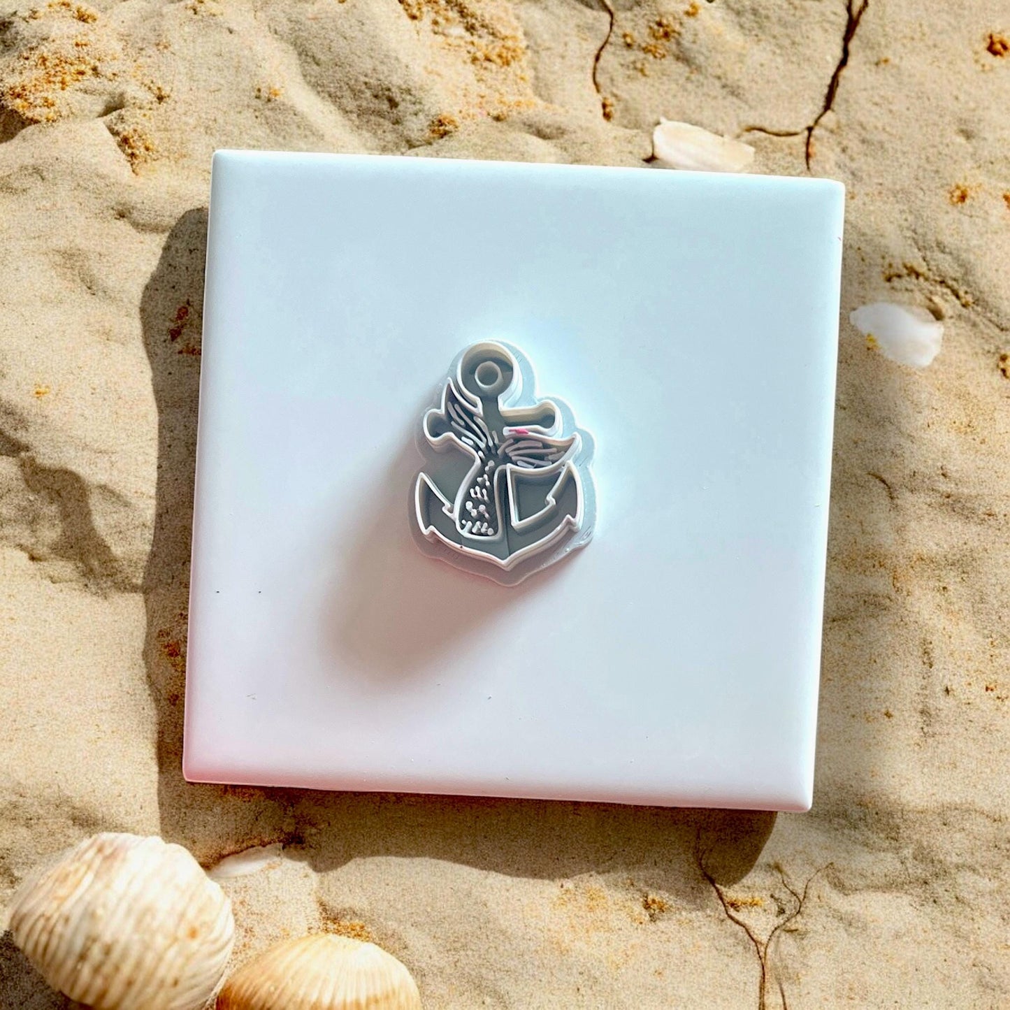 White polymer clay cutter in the shape of a mermaid tail anchor, resting on white tile with shells and sand.