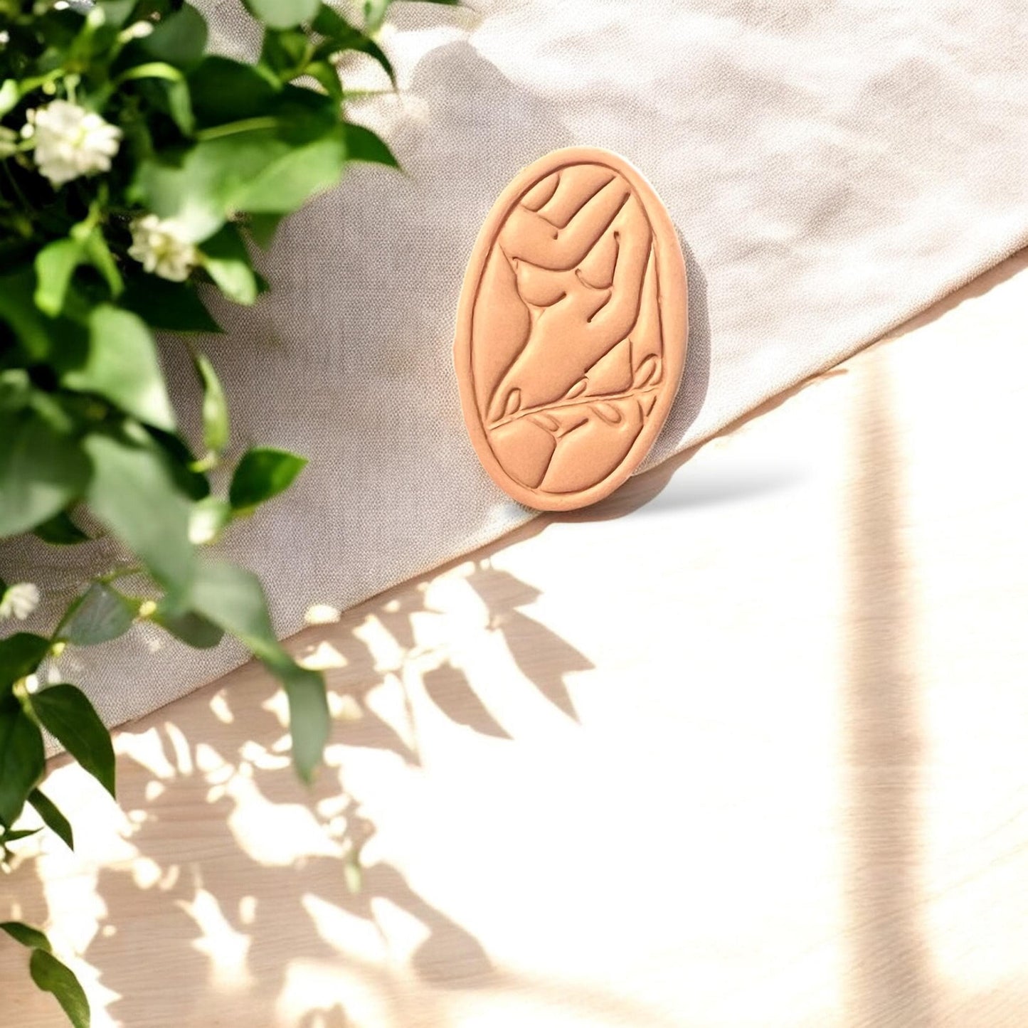 Nude clay cutout of feminine form resting on light linen with greenery in background, capturing the soft artistic vibe.