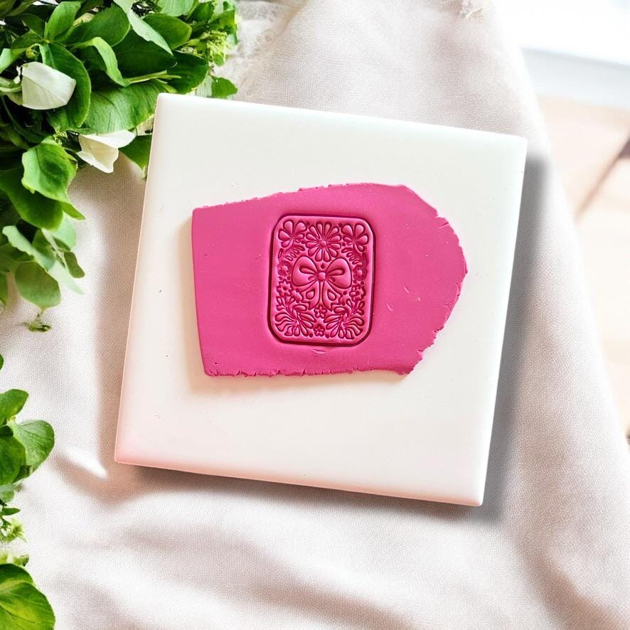 Coquette bow and floral motif clay imprint on pink polymer clay, displayed on white tile with greenery accents.