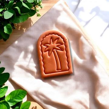 Orange polymer clay charm of a palm tree arch design, styled beautifully with soft fabric and fresh greenery.