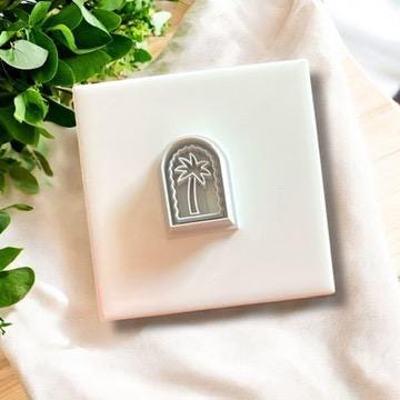 Gray palm tree arch clay cutter displayed on a white ceramic tile with greenery, perfect for coastal-themed jewelry.