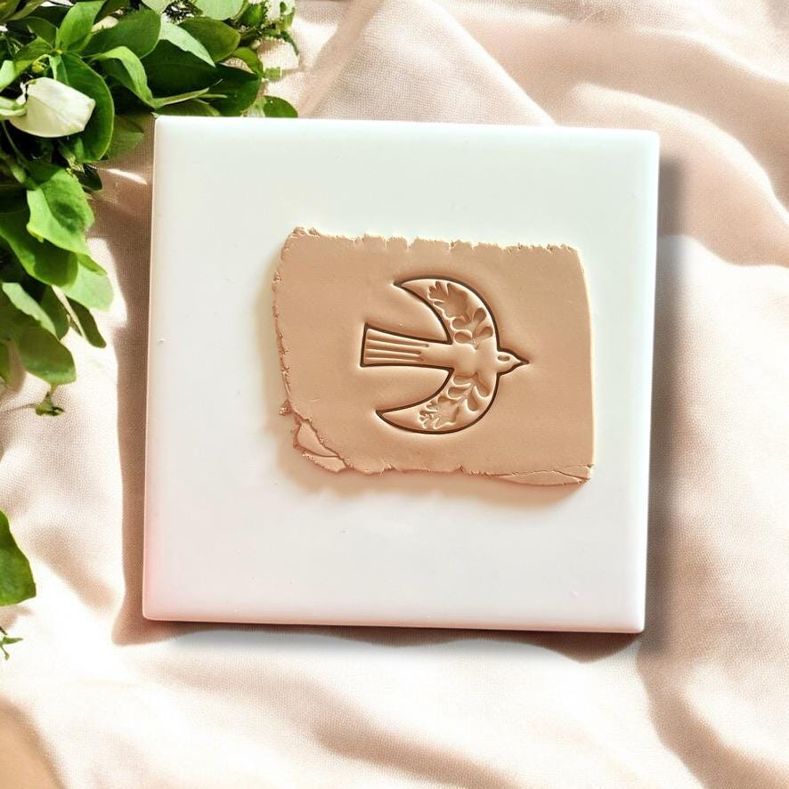 Boho Scandinavian-inspired folk bird clay imprint on beige polymer clay, displayed on a white tile with fresh greenery accents.