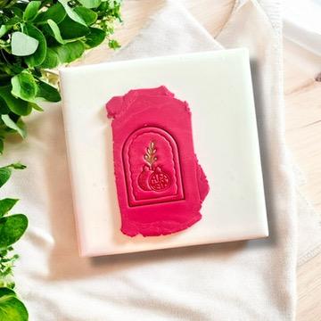Pomegranate arch clay imprint on pink polymer clay, displayed on a white tile with greenery accents.
