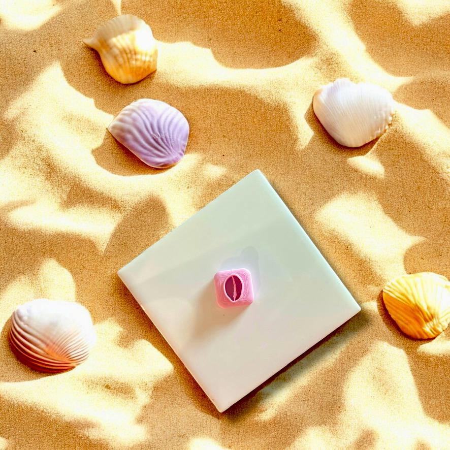 Pink cowrie shell clay cutter displayed on a white ceramic plate with beach sand and seashells for a coastal vibe.