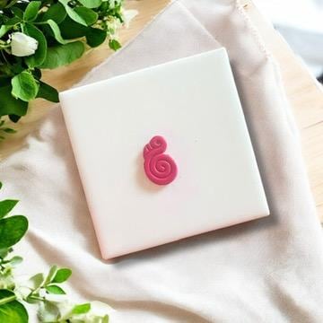 Small pink flamingo clay charm with a twisted design, displayed on a white ceramic tile with soft fabric.