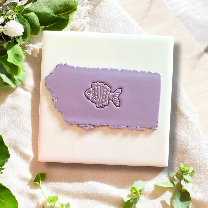 Detailed fish clay imprint on purple polymer clay, displayed on a white ceramic tile with fresh greenery accents.