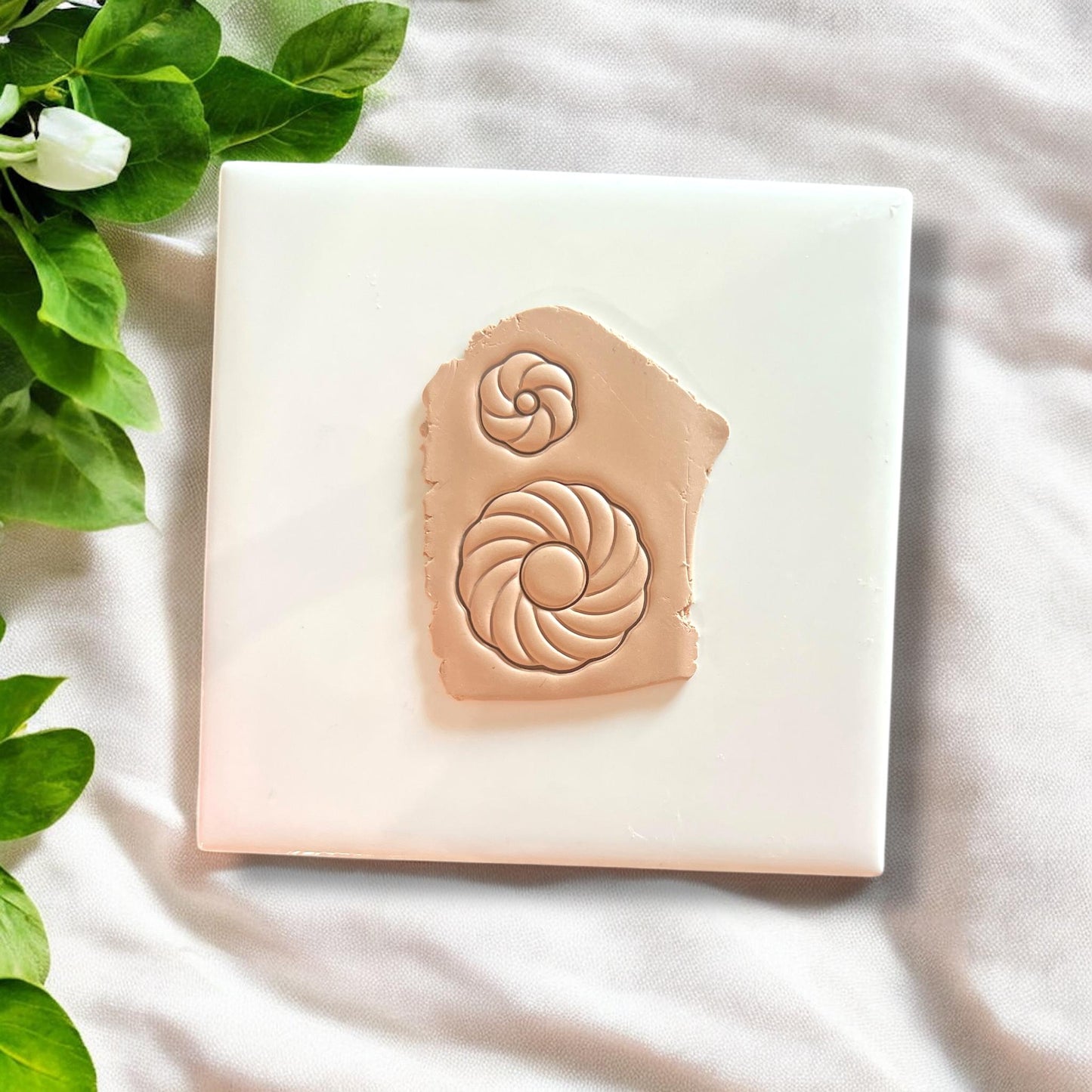 Polymer clay impression of swirl shell designs in tan clay on white tile with green leaves.