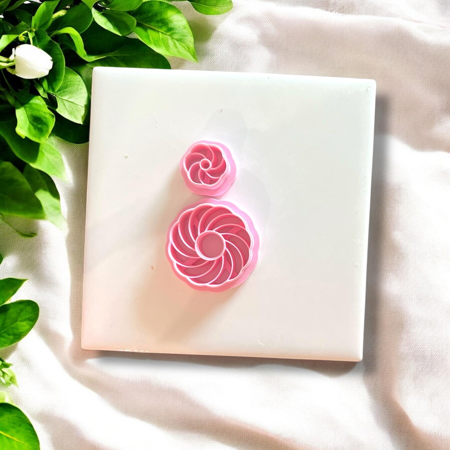 Pink polymer clay cutters in swirl shell designs, displayed on a white tile with green leaves.