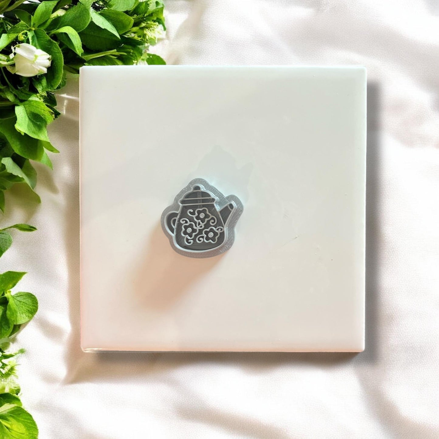 Floral teapot polymer clay cutter displayed alone on a white tile with leaves.