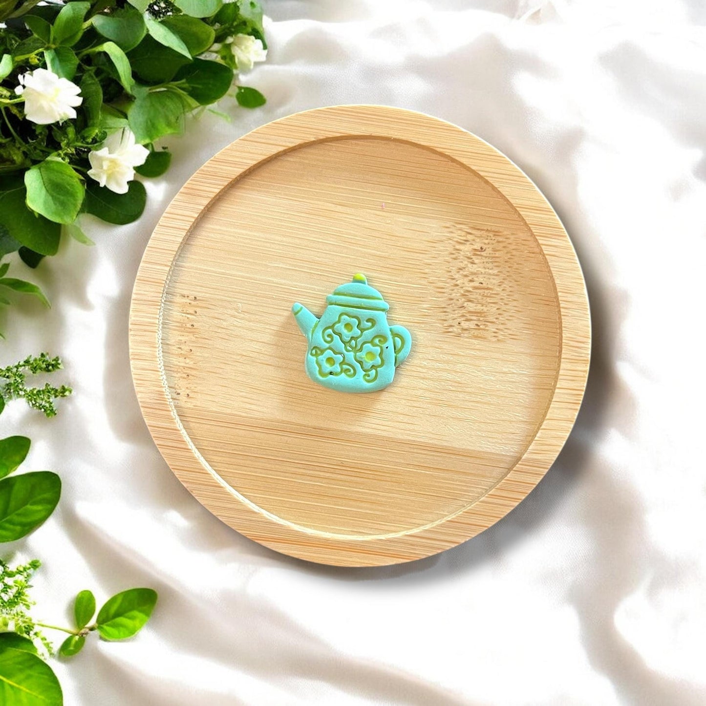 Finished teapot charm in light blue and green displayed on a bamboo tray, with floral detailing.