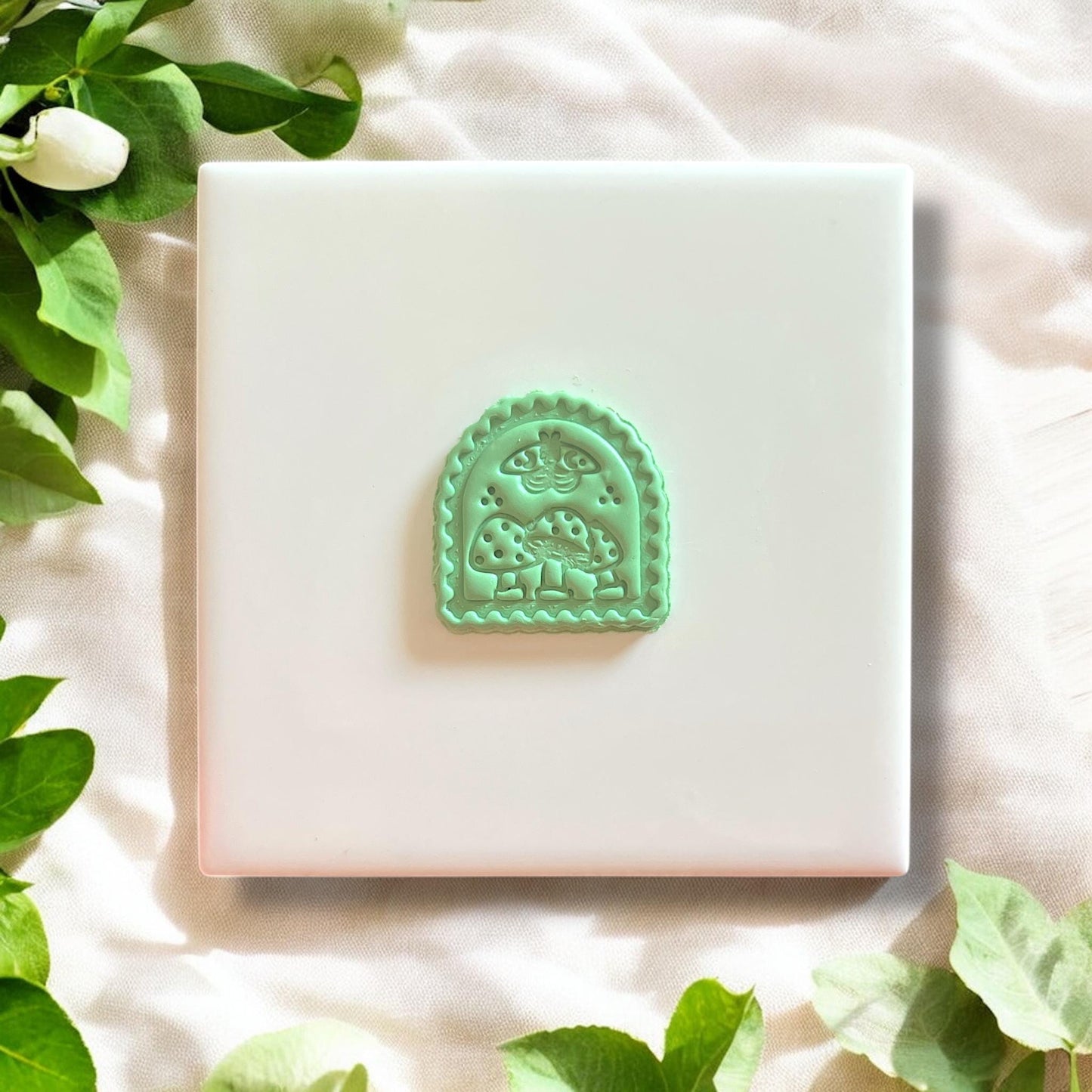 Mint green polymer clay impression of mushroom and moth design on a white tile with floral greenery.