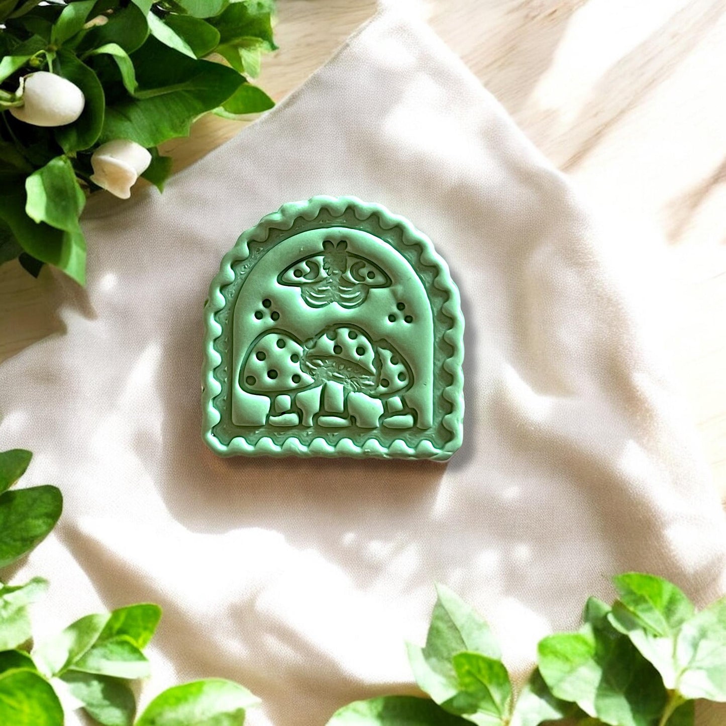 Mint green polymer clay charm with detailed mushroom and moth design shown on soft fabric with floral leaves.