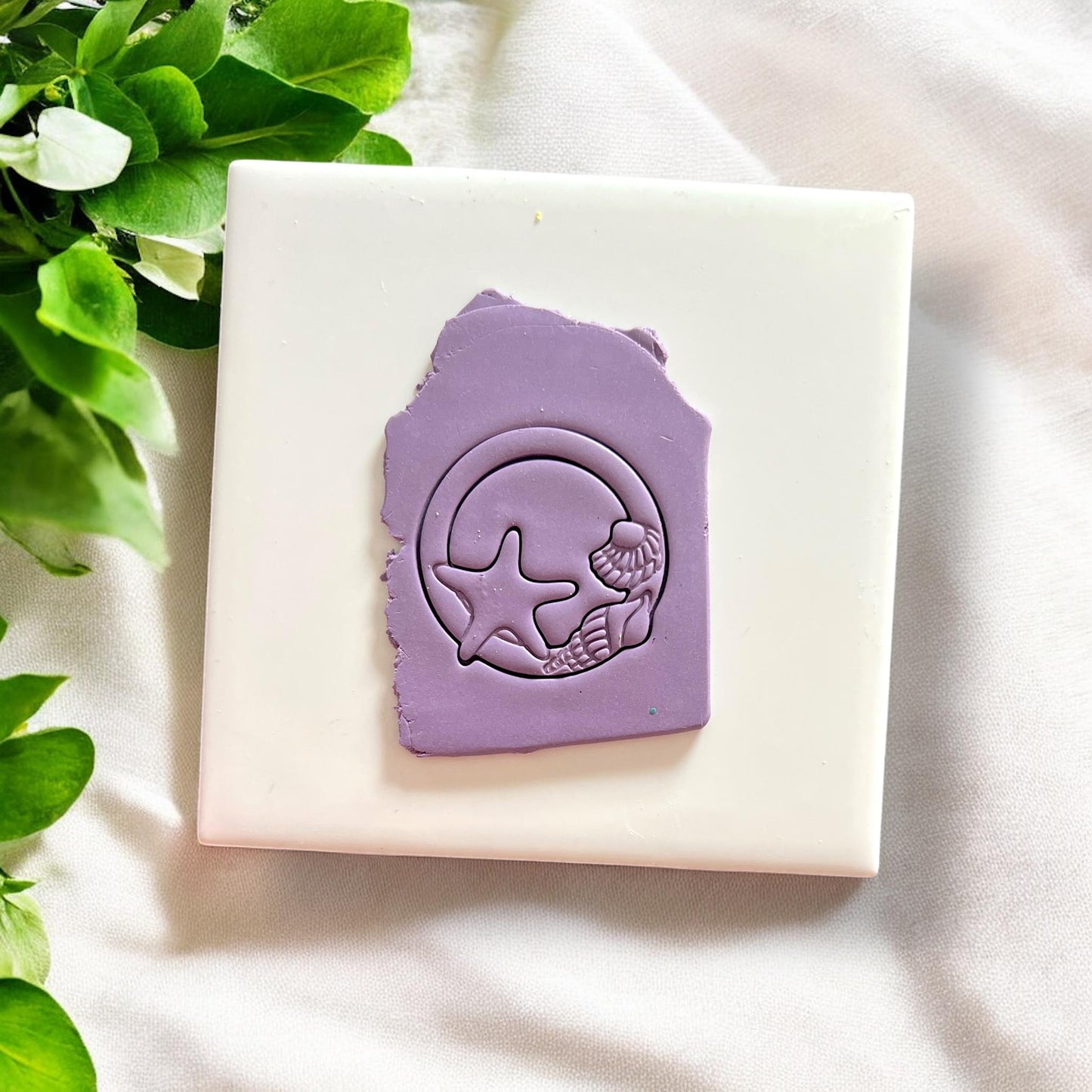 Purple polymer clay slab stamped with seashell circle cutter showing starfish and shell details on a white tile