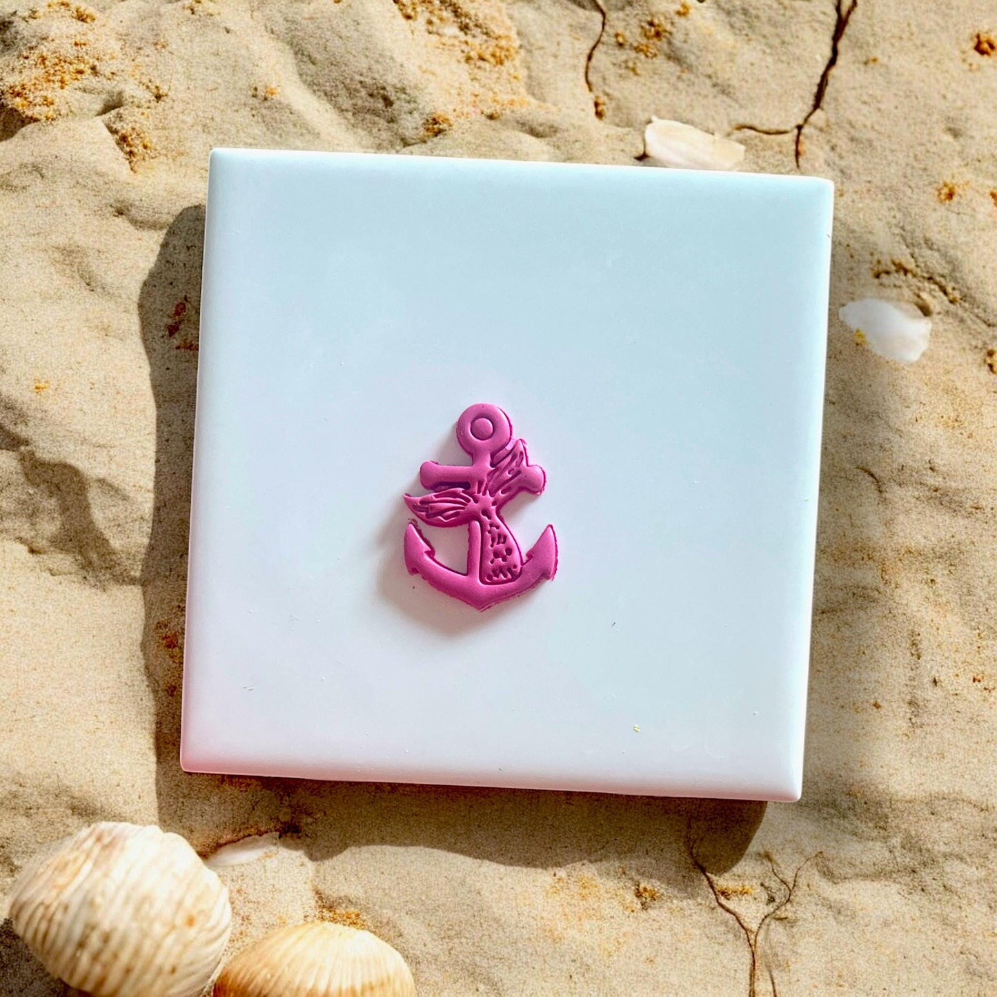 Finished polymer clay anchor with mermaid tail design on white tile with seashells and sand.