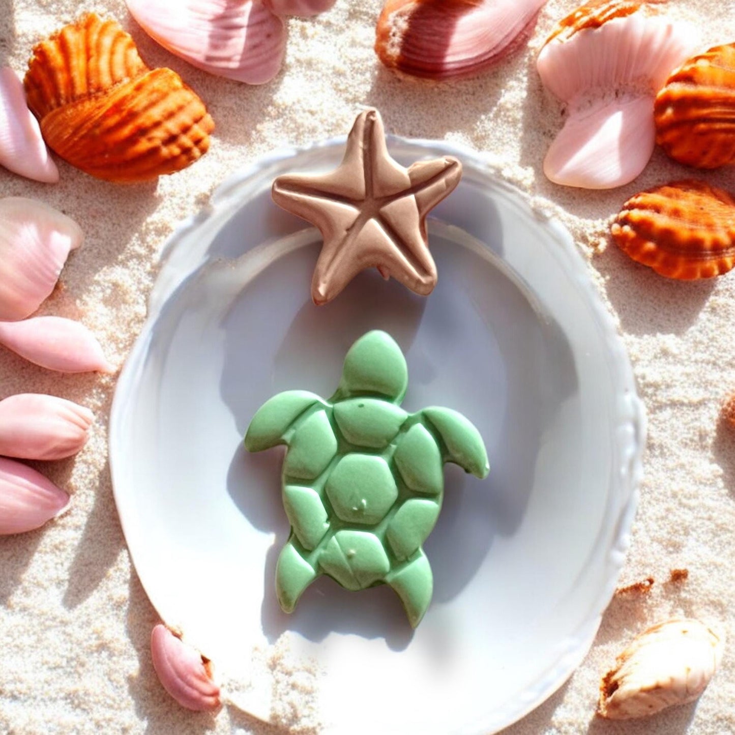 Green turtle and bronze starfish clay earrings displayed on a seashell dish in beach sand