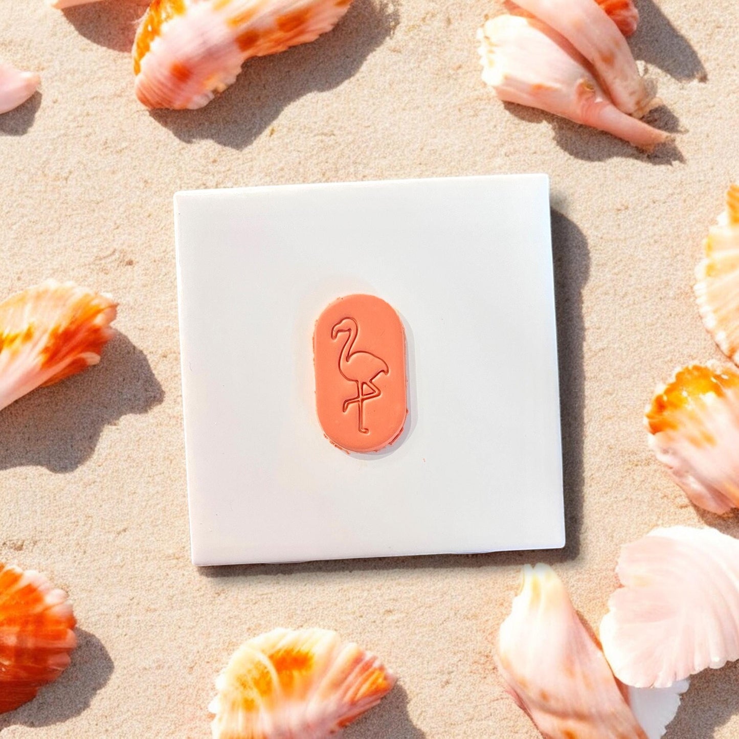 Finished orange flamingo clay piece on white square background, tropical shell decor surrounding it