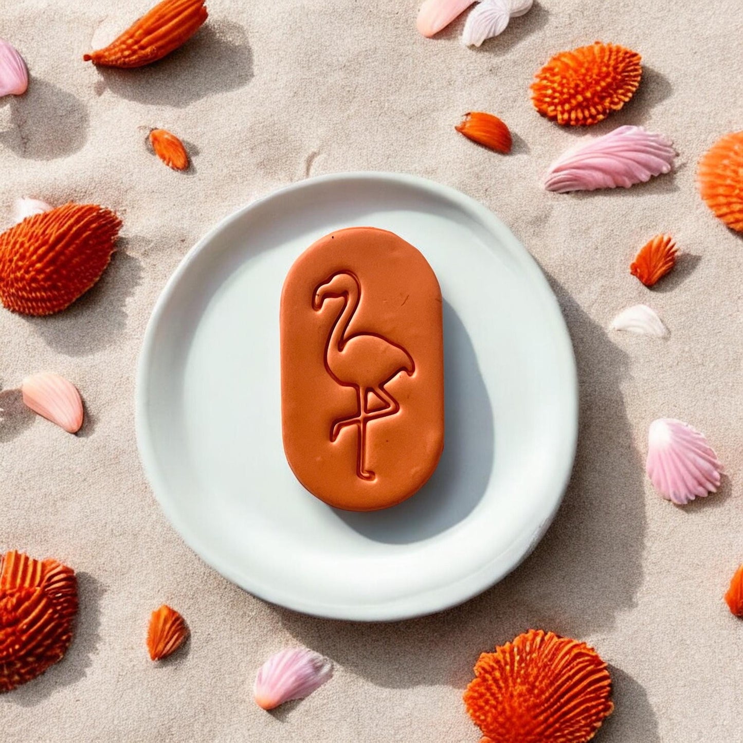 Close-up of full orange flamingo clay piece on a white dish, styled on sand with seashells around