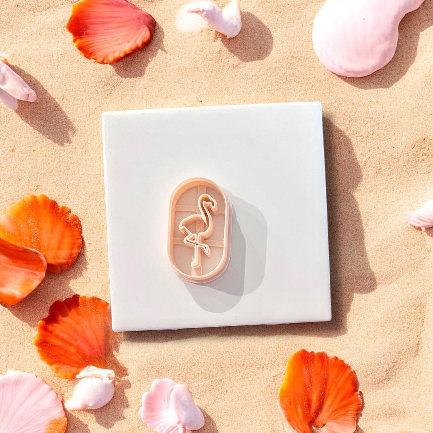Top view of oval flamingo cutter placed on white tile, surrounded by beach-themed orange and pink shells