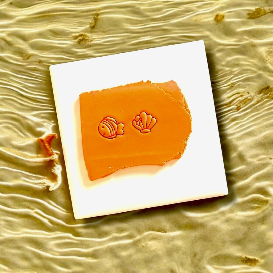 Fish and seashell-shaped clay cutter imprints on orange polymer clay, displayed on white tile with ocean wave background.