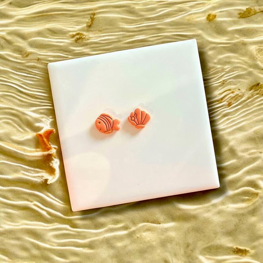 Orange fish and shell clay charms made from polymer clay, displayed on a white tile with beach-inspired background.