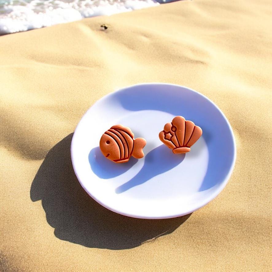 Fish and seashell polymer clay charms displayed on a white plate set against beach sand and ocean waves in the background.