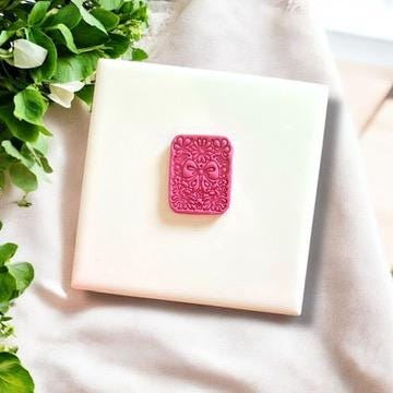 Small pink clay charm with coquette bow and floral design, displayed on a white ceramic tile surrounded by fresh greenery.