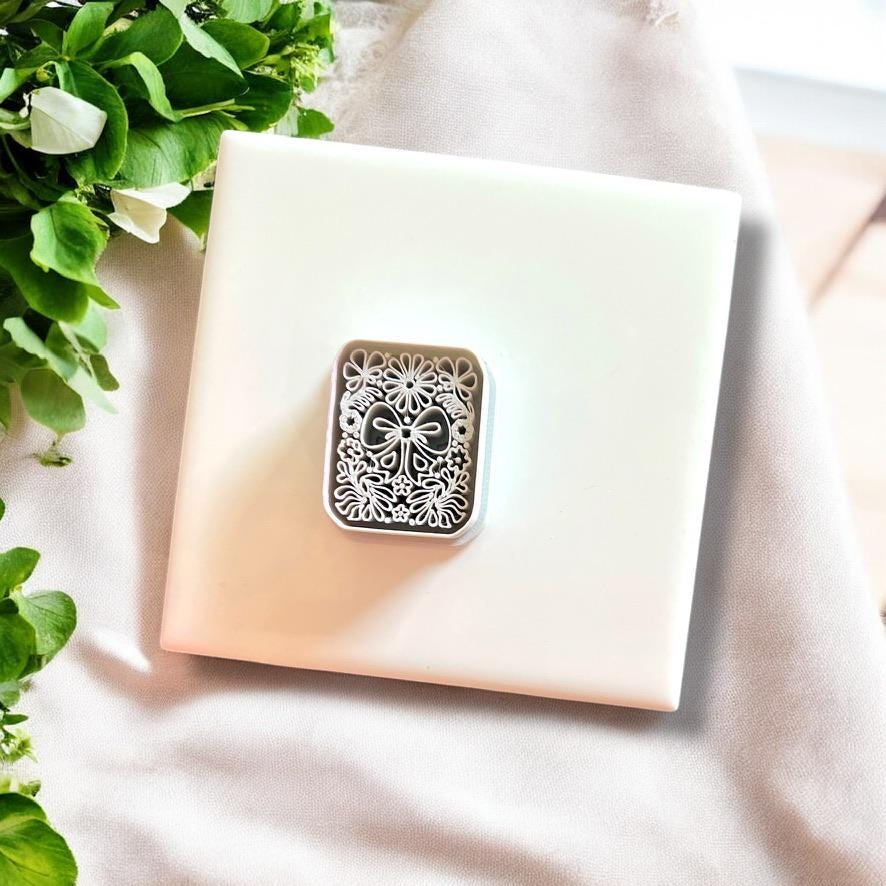 Large coquette bow and floral motif imprint on pink polymer clay, styled beautifully with greenery on a soft fabric backdrop.