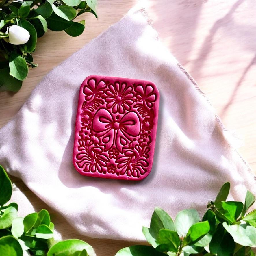 Small pink clay charm with coquette bow and floral design, displayed on a white ceramic tile surrounded by fresh greenery.
