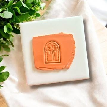 Palm tree arch clay imprint on orange polymer clay, displayed on a white ceramic tile with greenery accents.
