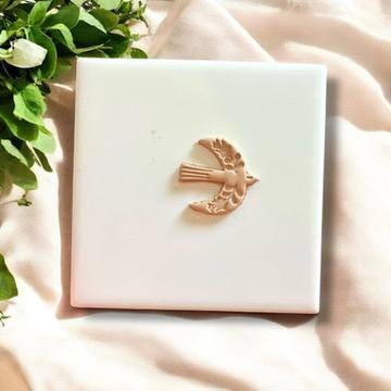 Beige polymer clay charm with a Scandinavian folk bird design, displayed on a white tile surrounded by greenery.