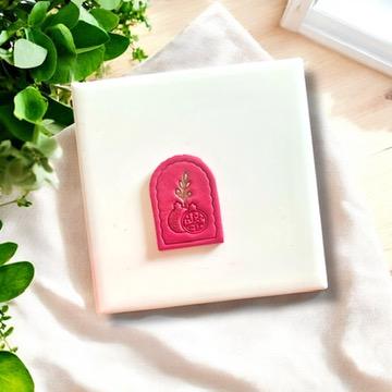 Pink clay charm with a pomegranate arch design, displayed on a white ceramic tile with soft greenery in the background.