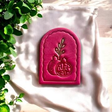 Large polymer clay pomegranate arch design in pink, beautifully displayed on soft fabric with greenery accents.
