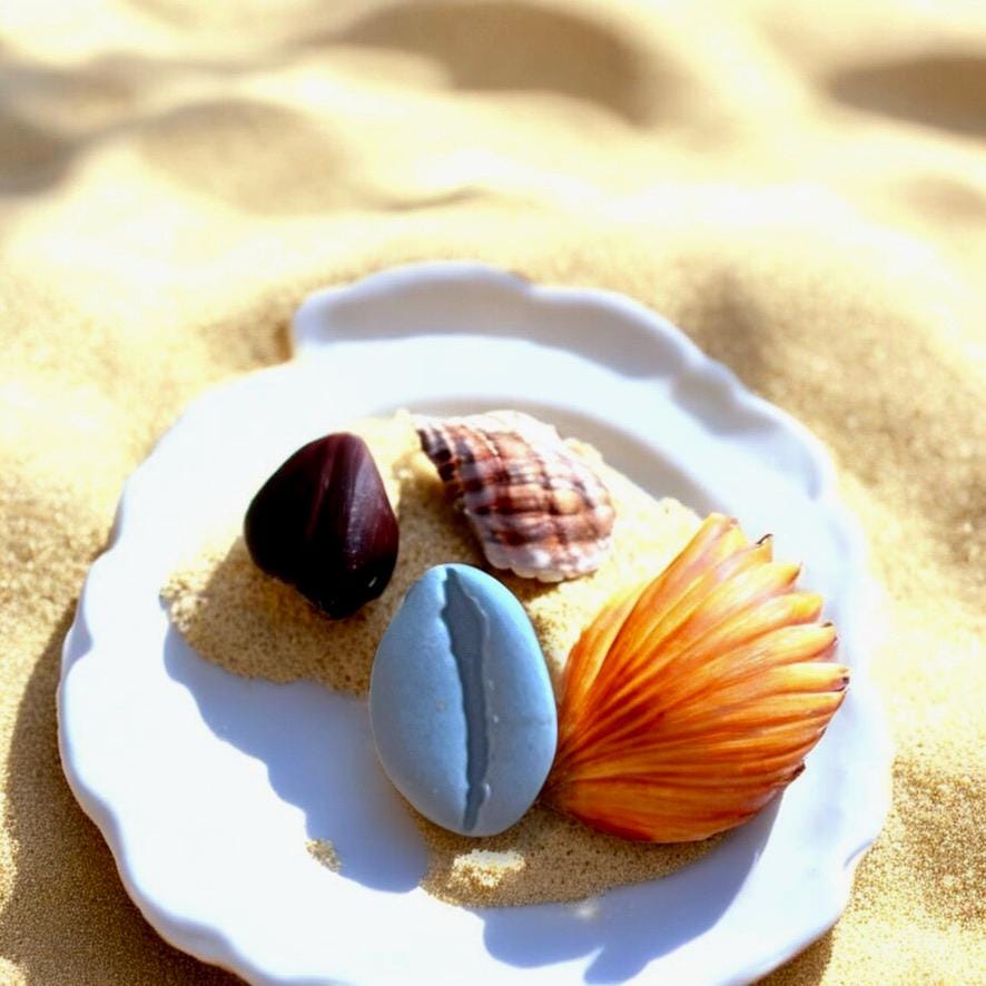 Blue polymer clay cowrie shell charm displayed with real seashells on a white plate filled with beach sand.