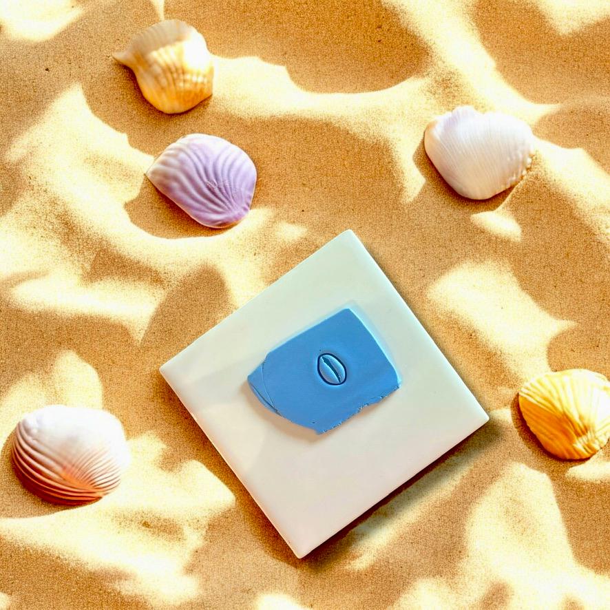 Cowrie shell clay imprint on blue polymer clay, displayed on white tile with seashells and beach sand accents.