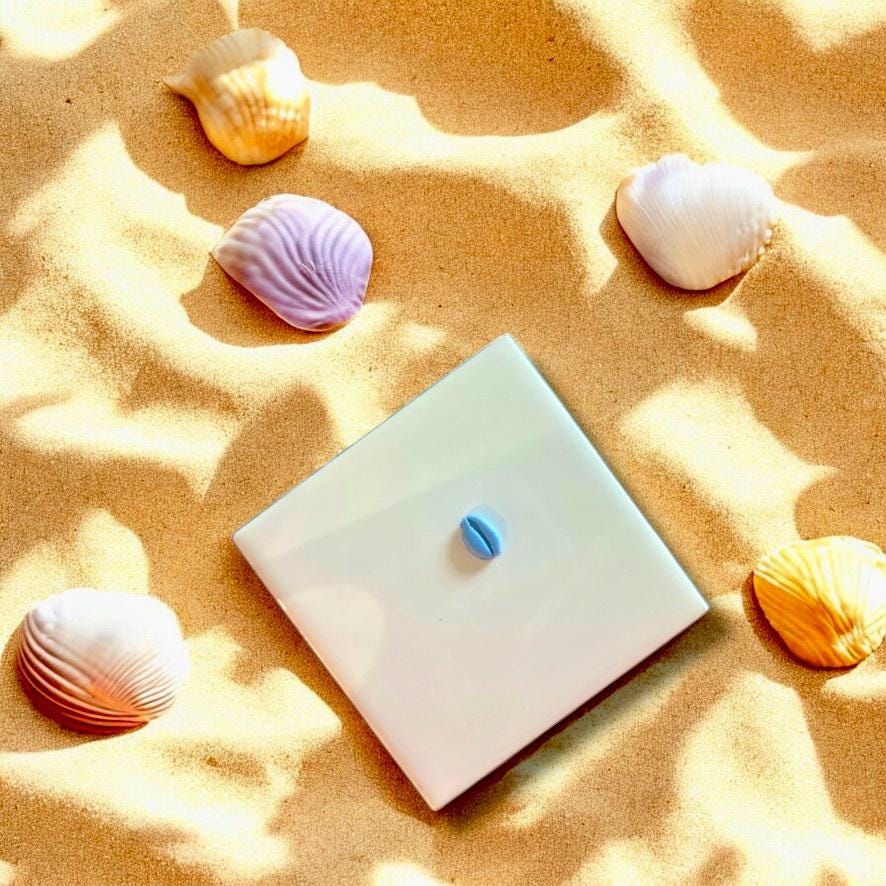 Blue polymer clay cowrie shell charm displayed on a white ceramic tile surrounded by sand and shells.