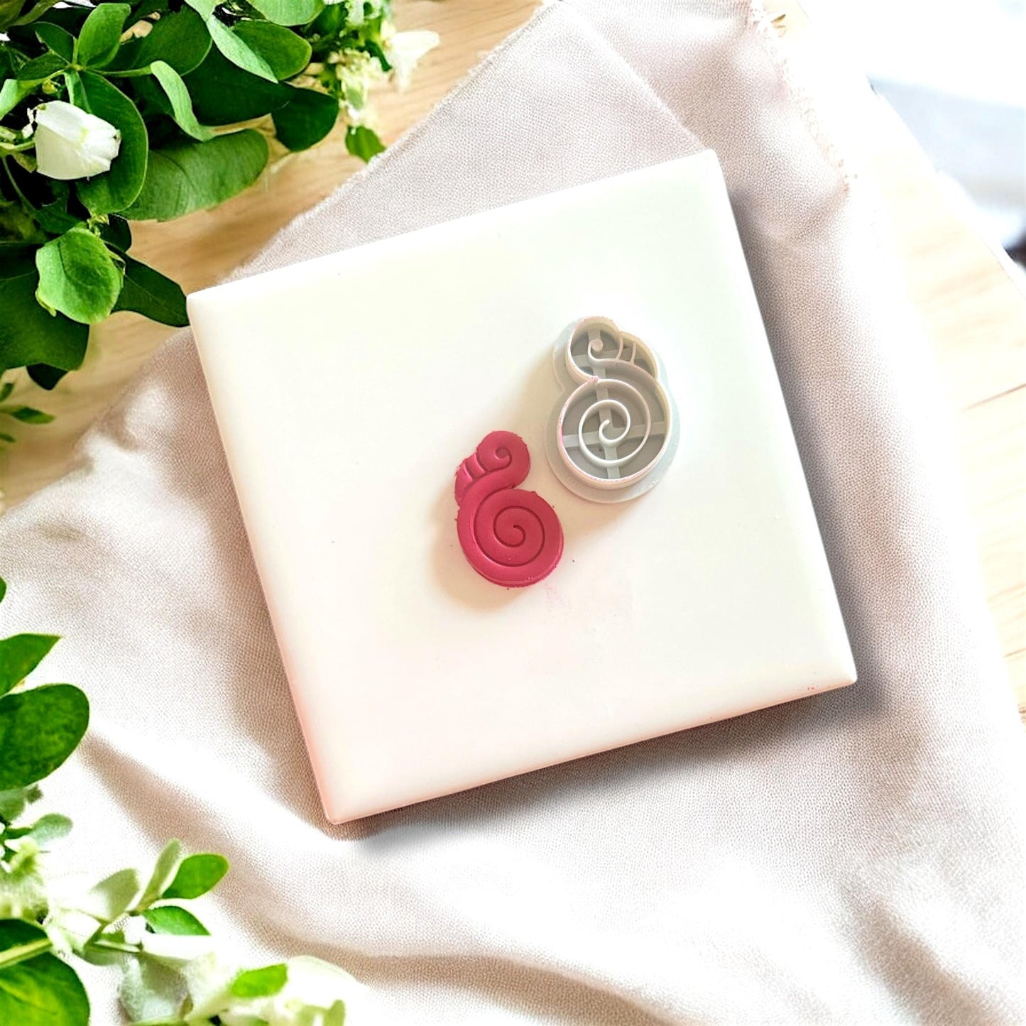 Flamingo clay charm alongside its matching cutter, displayed on a white tile with fresh greenery and a fabric backdrop.