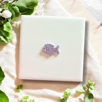 Purple polymer clay fish charm with intricate details, displayed on a white tile with soft green leaves.