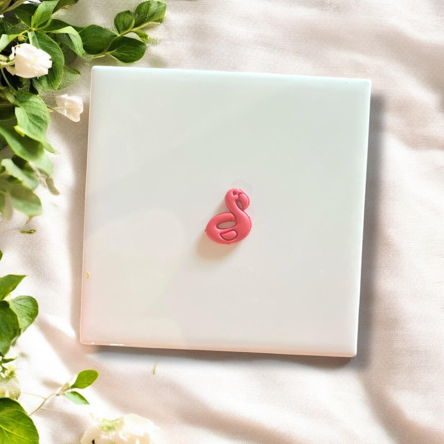 Finished polymer clay flamingo floatie charm displayed on white tile with greenery accents.