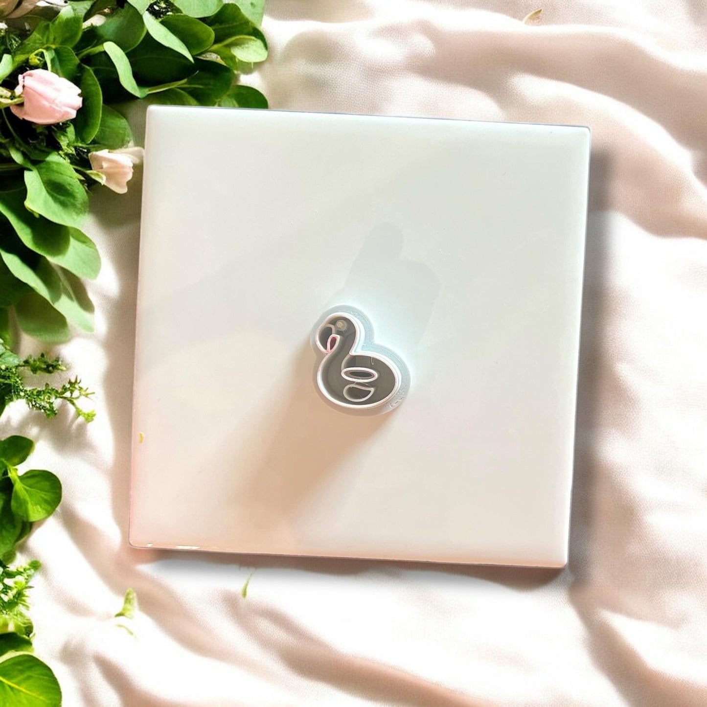 White flamingo floatie polymer clay cutter displayed on white tile with greenery.