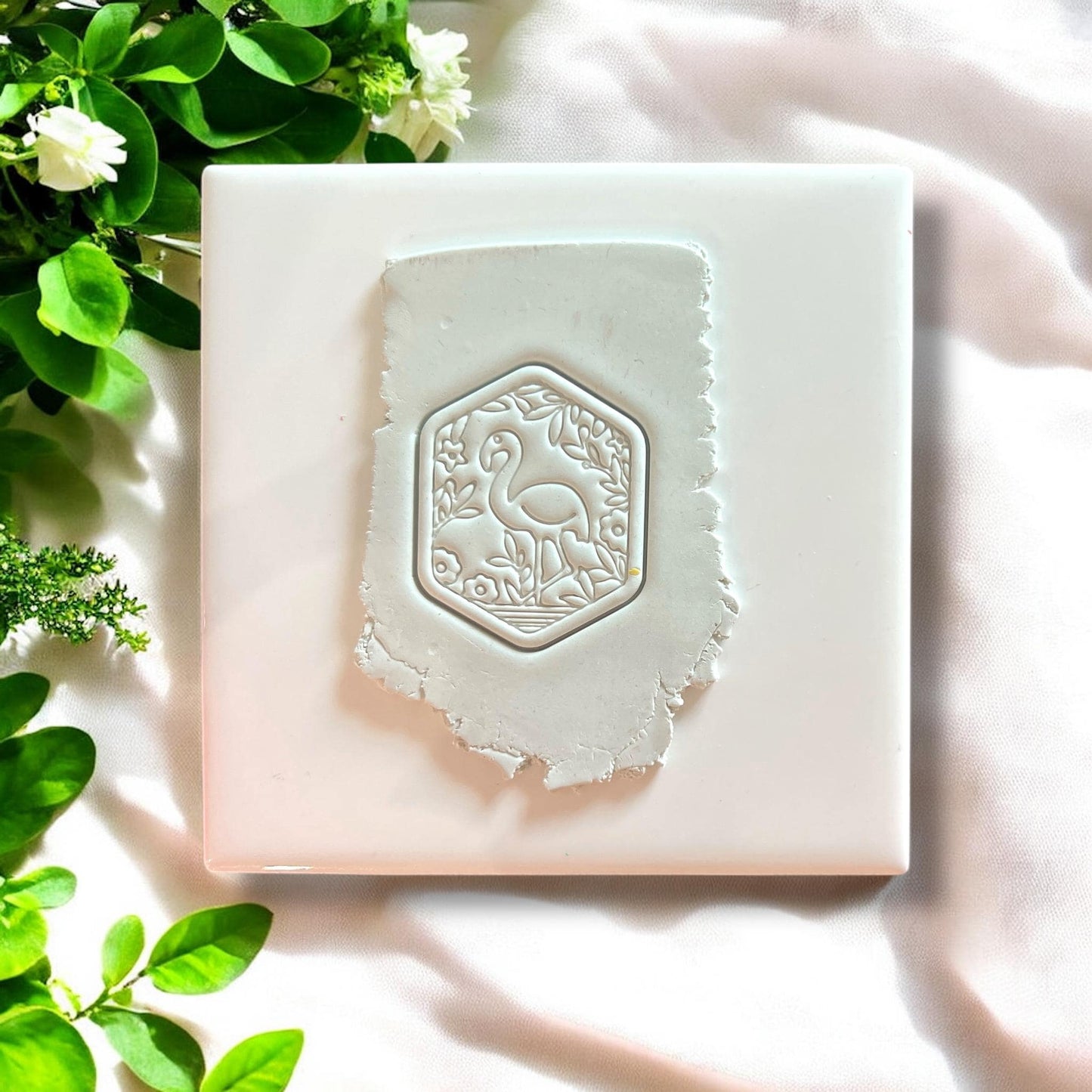 Polymer clay impression of a garden flamingo design in white clay on a white tile with greenery.