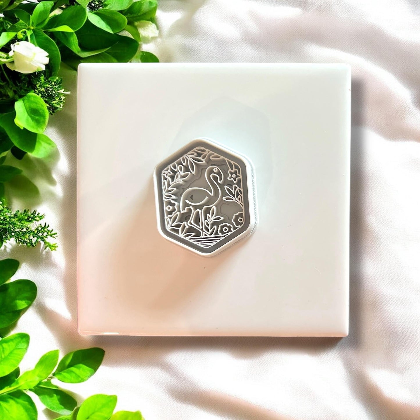 White hexagon-shaped cutter with intricate garden flamingo design on a white tile with green leaves.
