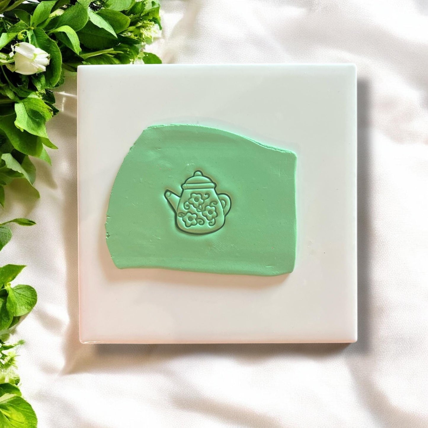 Green polymer clay impression of a floral teapot design on a white tile with greenery accents.