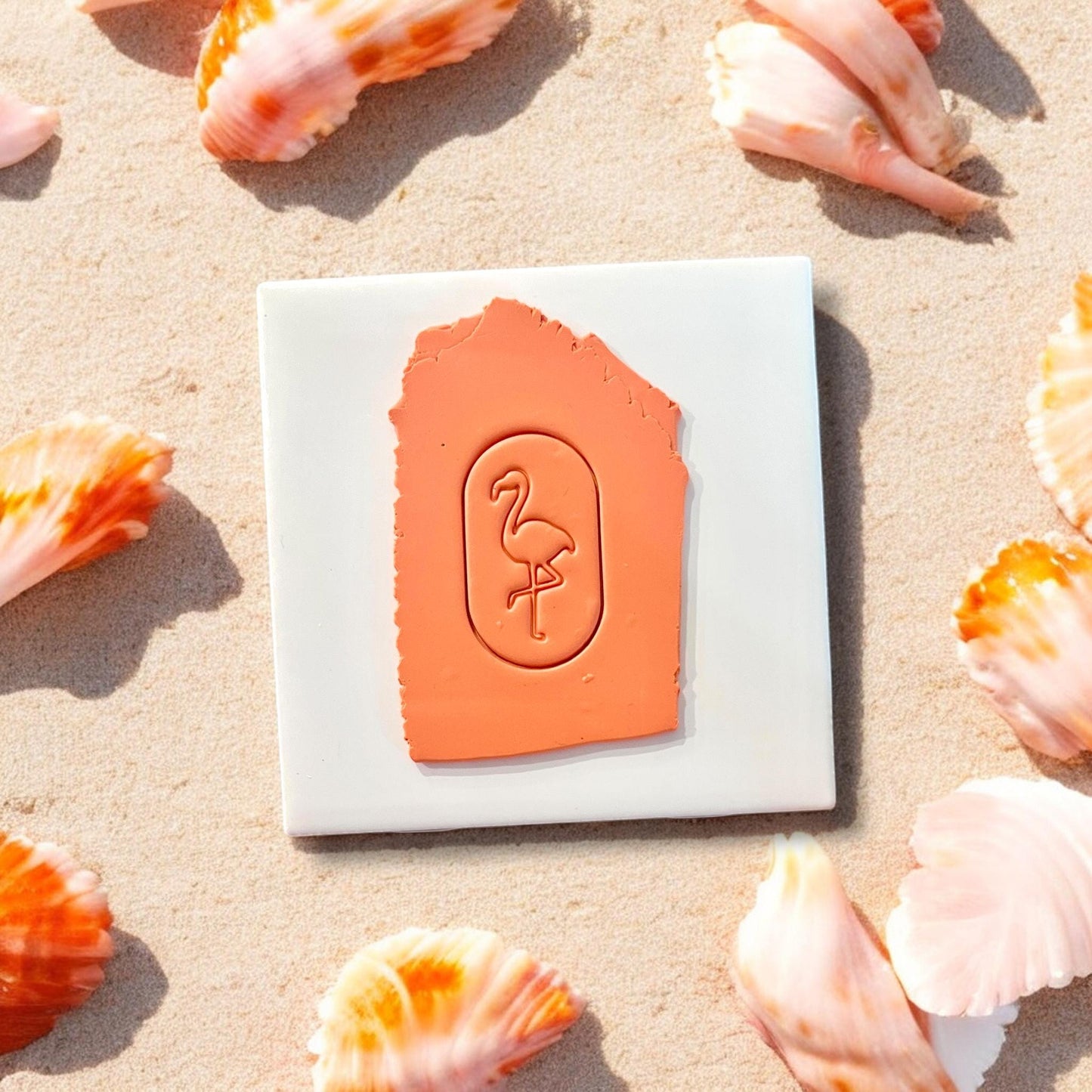 Oval flamingo cutter imprint in orange clay on white tile, surrounded by seashells on beach sand