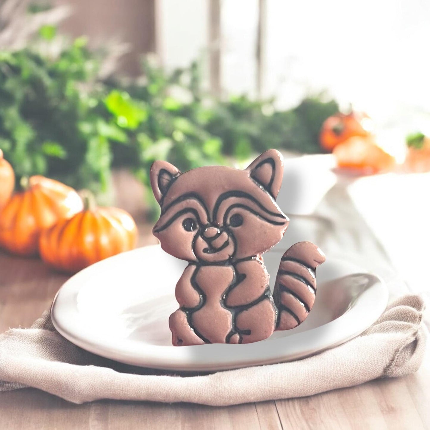 Standing raccoon clay earring displayed upright on white plate with autumn props