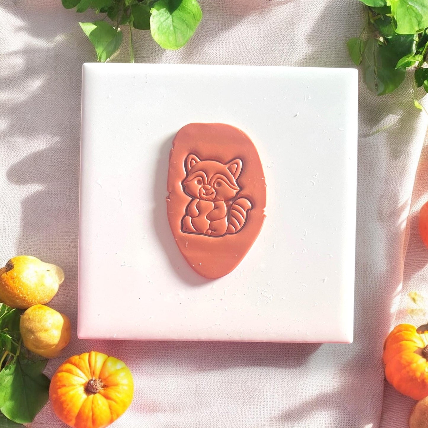 Raccoon clay shape stamped into light clay on white tile with pumpkins around